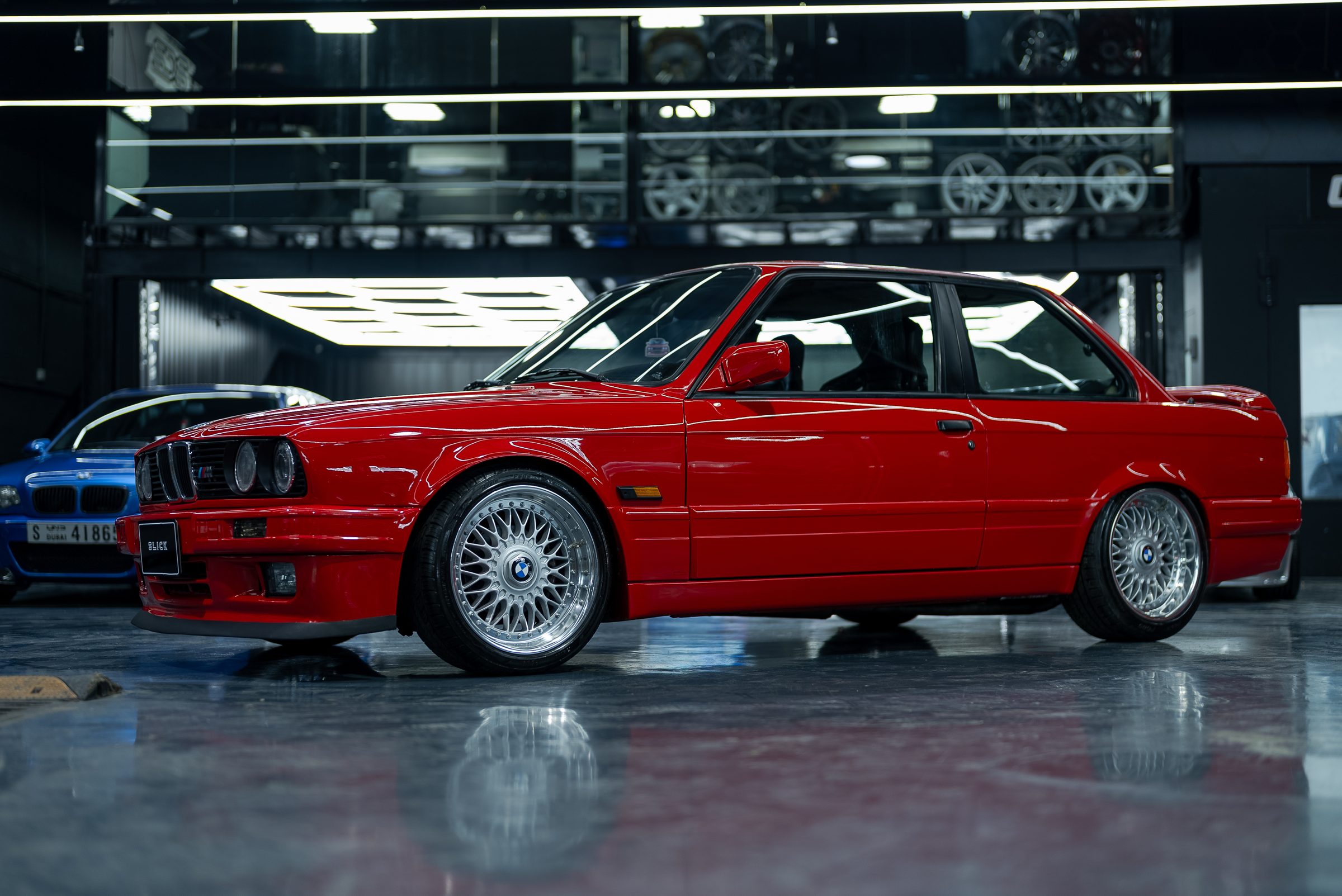 A red BMW E30 M3 on custom wheels inside the workshop